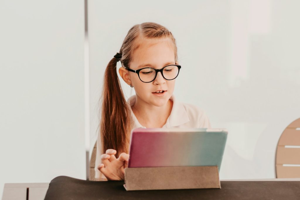 Teenage girl wearing eyeglasses and white polo shirt sitting at table with tablet pad in videochat